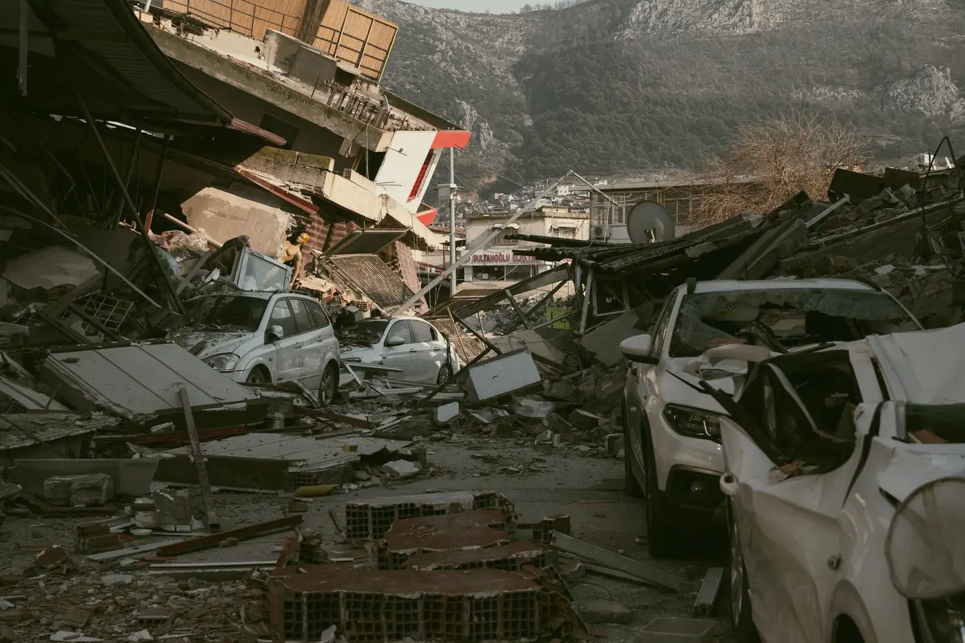 A street of collapsed buildings and crushed cars buried in rubble after an earthquake, with hills in the background.