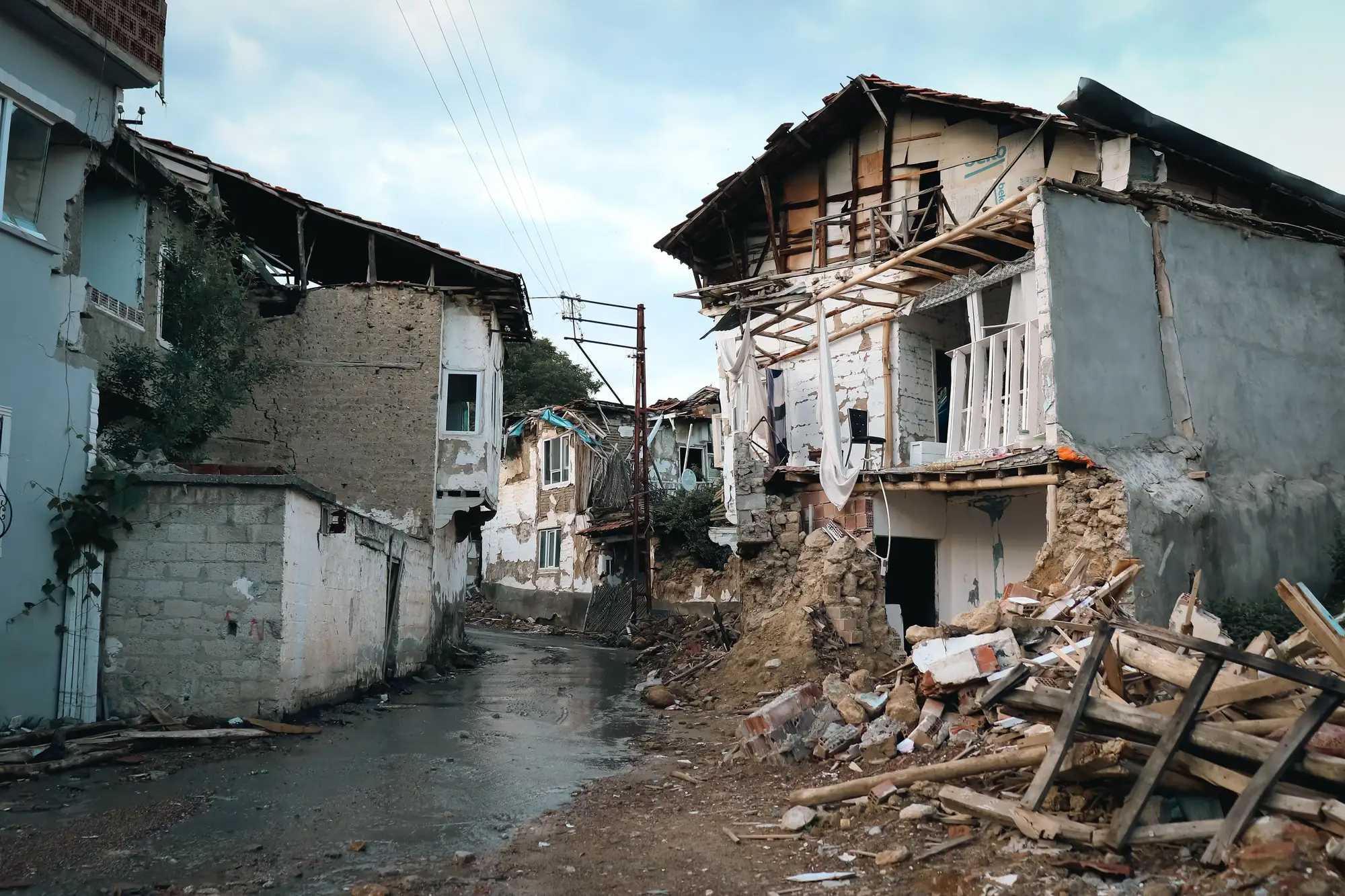 A street of earthquake-damaged houses: collapsed walls, broken roofs and timber debris across the road